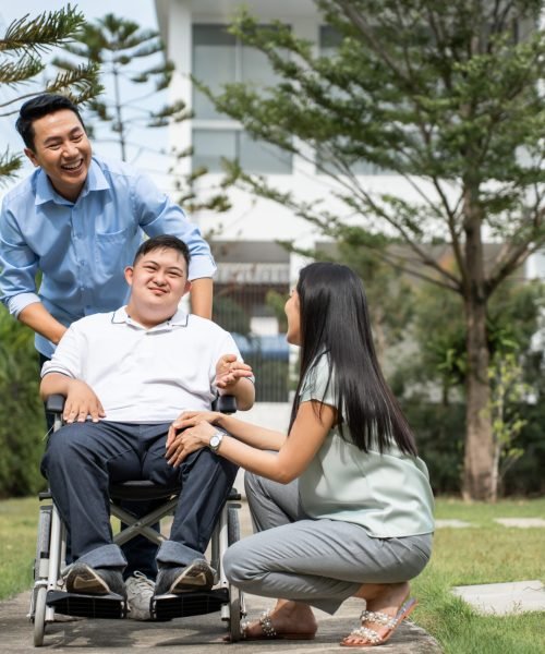 Asian attractive family walking with young son on wheelchair in garden. Beautiful loving couple, father and mother support take care young man people on wheelchair outdoors in the backyard at home.