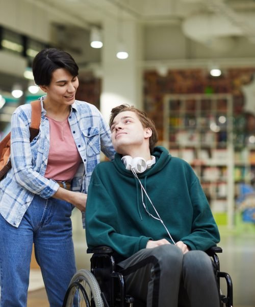 Positive attractive young woman with satchel pushing wheelchair and talking to disabled student in library